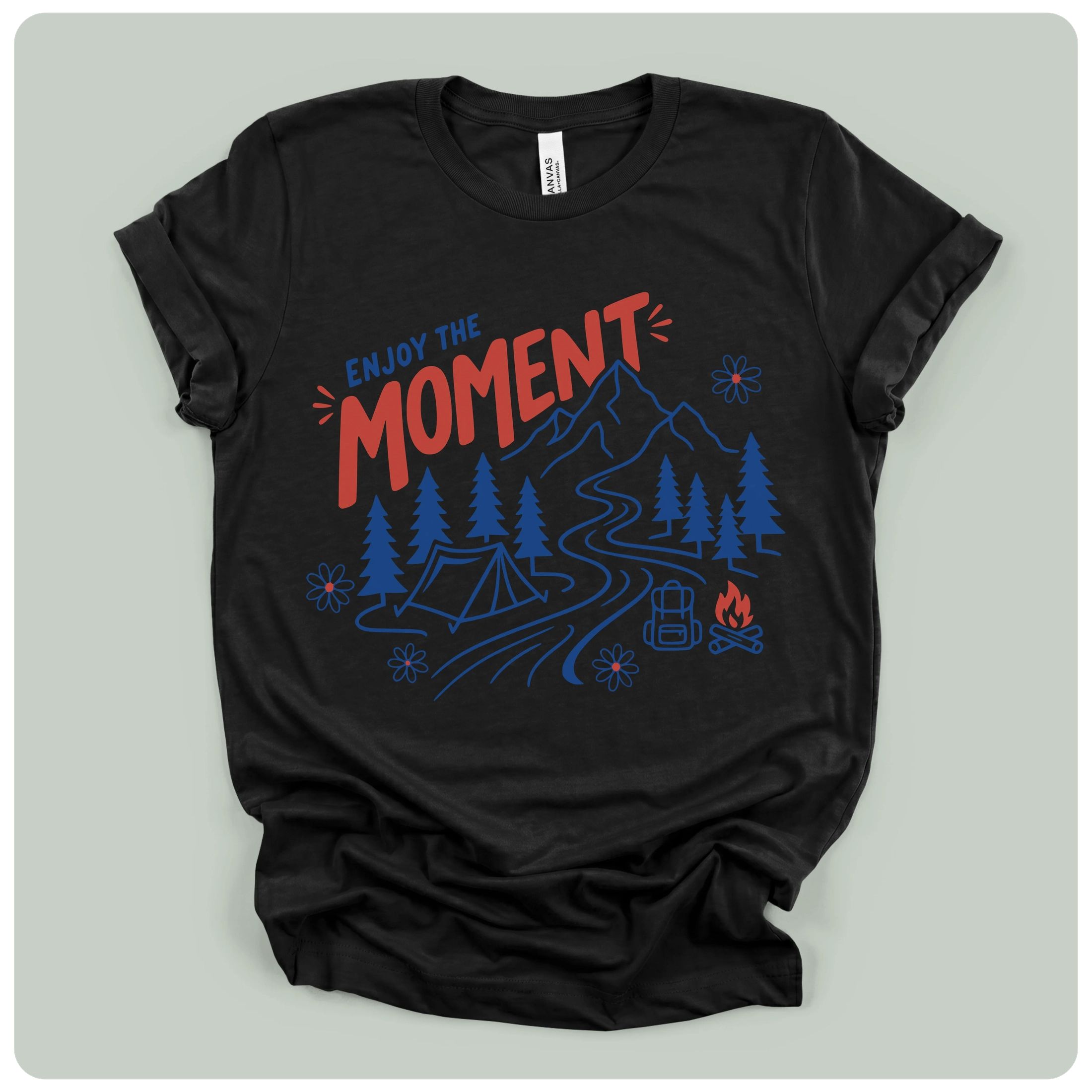 A black short-sleeve t-shirt with a graphic print on the front, laid flat on a light gray background. The graphic features the words 'ENJOY THE MOMENT' in red and blue, above a blue camping scene with mountains, trees, a tent, a river, and a campfire.