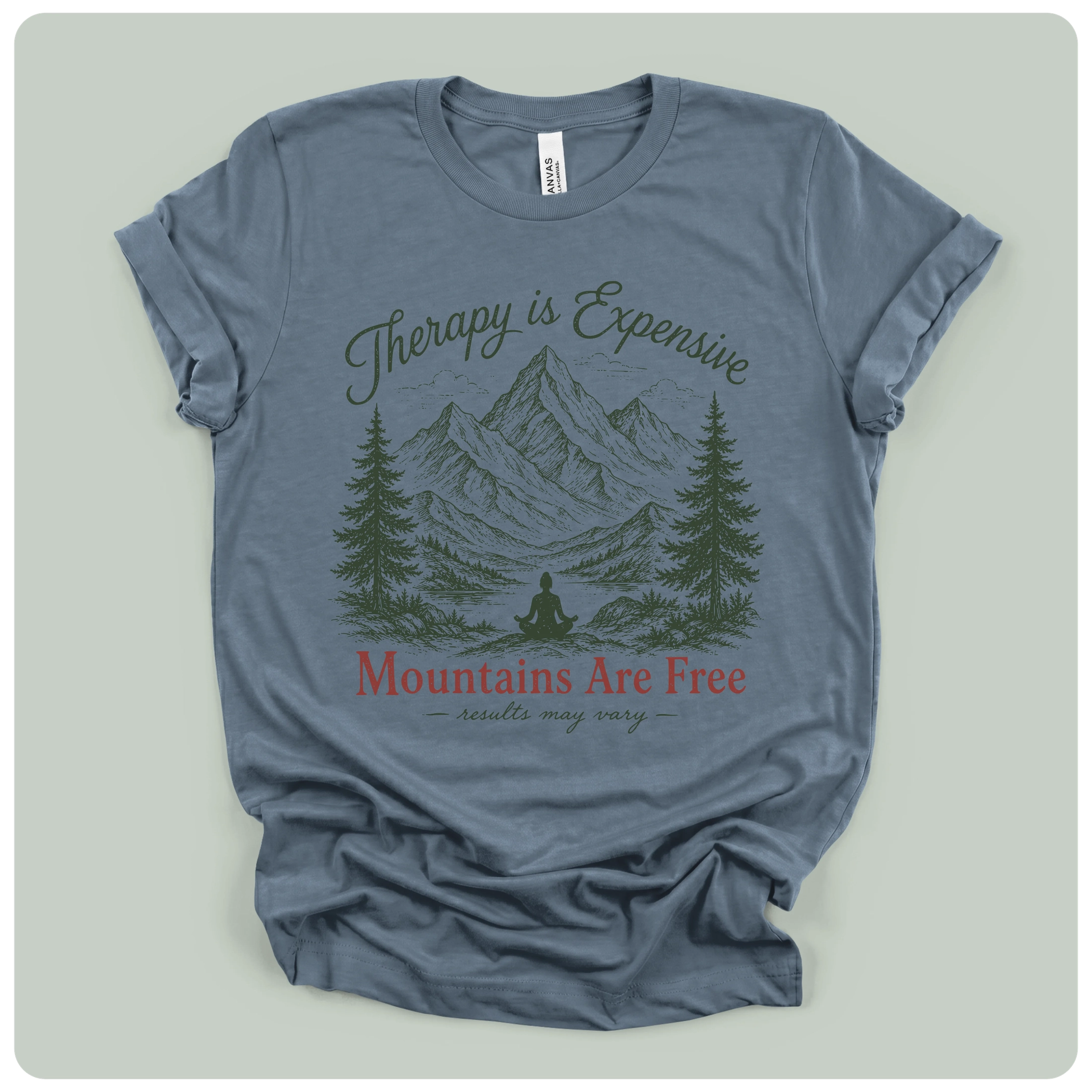 A slate blue crewneck t-shirt with a graphic print of mountains, pine trees, and a meditating figure. The text reads 'Therapy is Expensive' in dark green script, 'Mountains Are Free' in red serif font, and 'adjustments may vary' in small dark green script below.