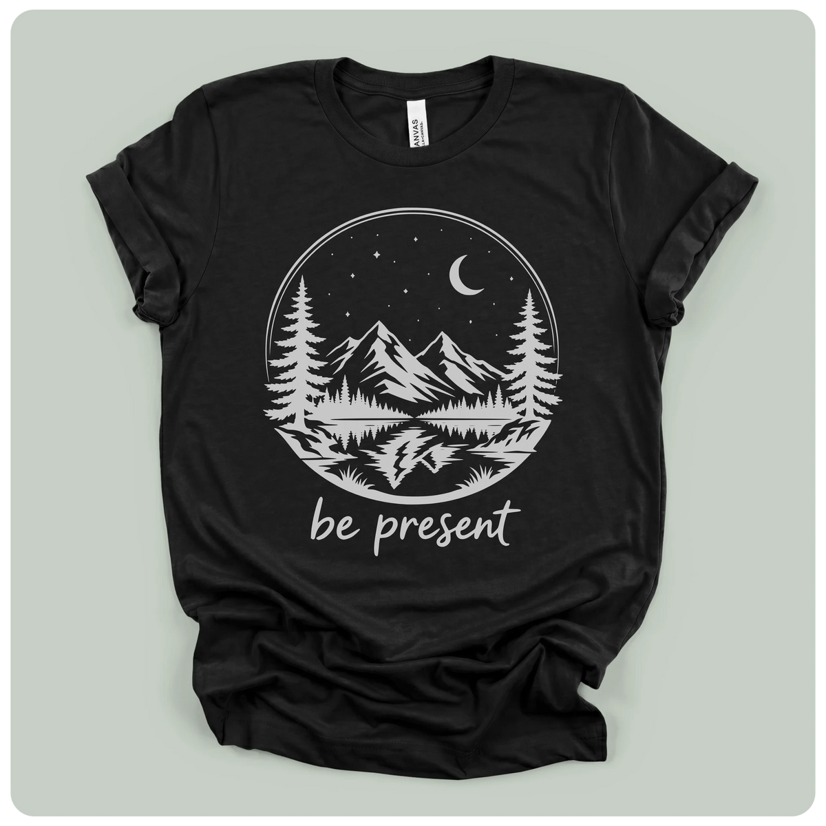 A black short-sleeve t-shirt with a white circular graphic of mountains, pine trees, a crescent moon, and stars, with the text 'be present' printed below. The t-shirt is laid flat on a light gray background.
