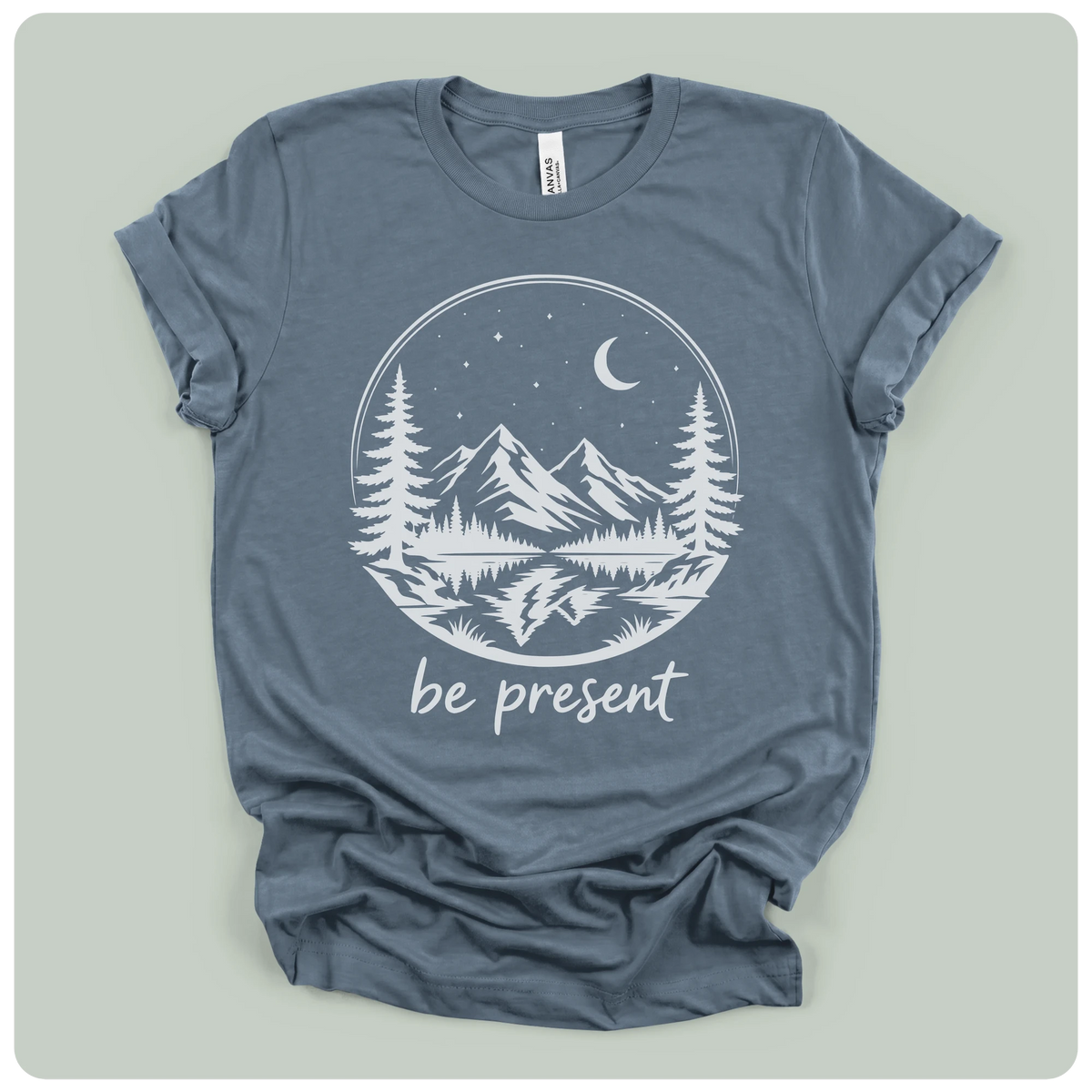 A slate blue crewneck t-shirt with a white graphic print of a mountain landscape at night with the text 'be present' underneath, laid flat on a light gray background.
