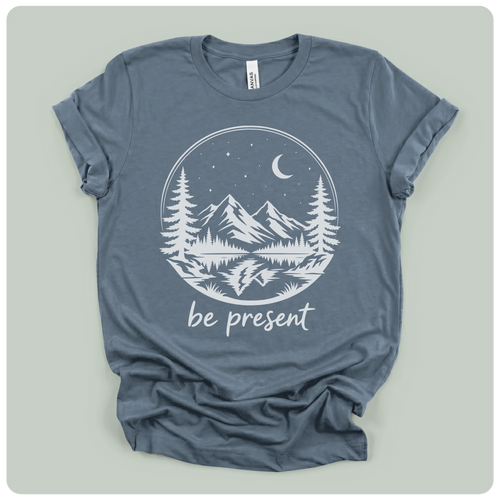 A slate blue crewneck t-shirt with a white graphic print of a mountain landscape at night with the text 'be present' underneath, laid flat on a light gray background.