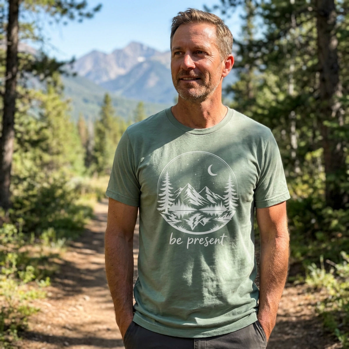 A man standing on a forest path wearing a sage green t-shirt with a white graphic of mountains, trees, and a crescent moon with the text 'be present'.