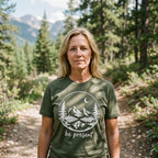 A woman standing on a forest path wearing an olive green t-shirt with a white graphic of mountains, trees, a crescent moon, and the text 'be present'.