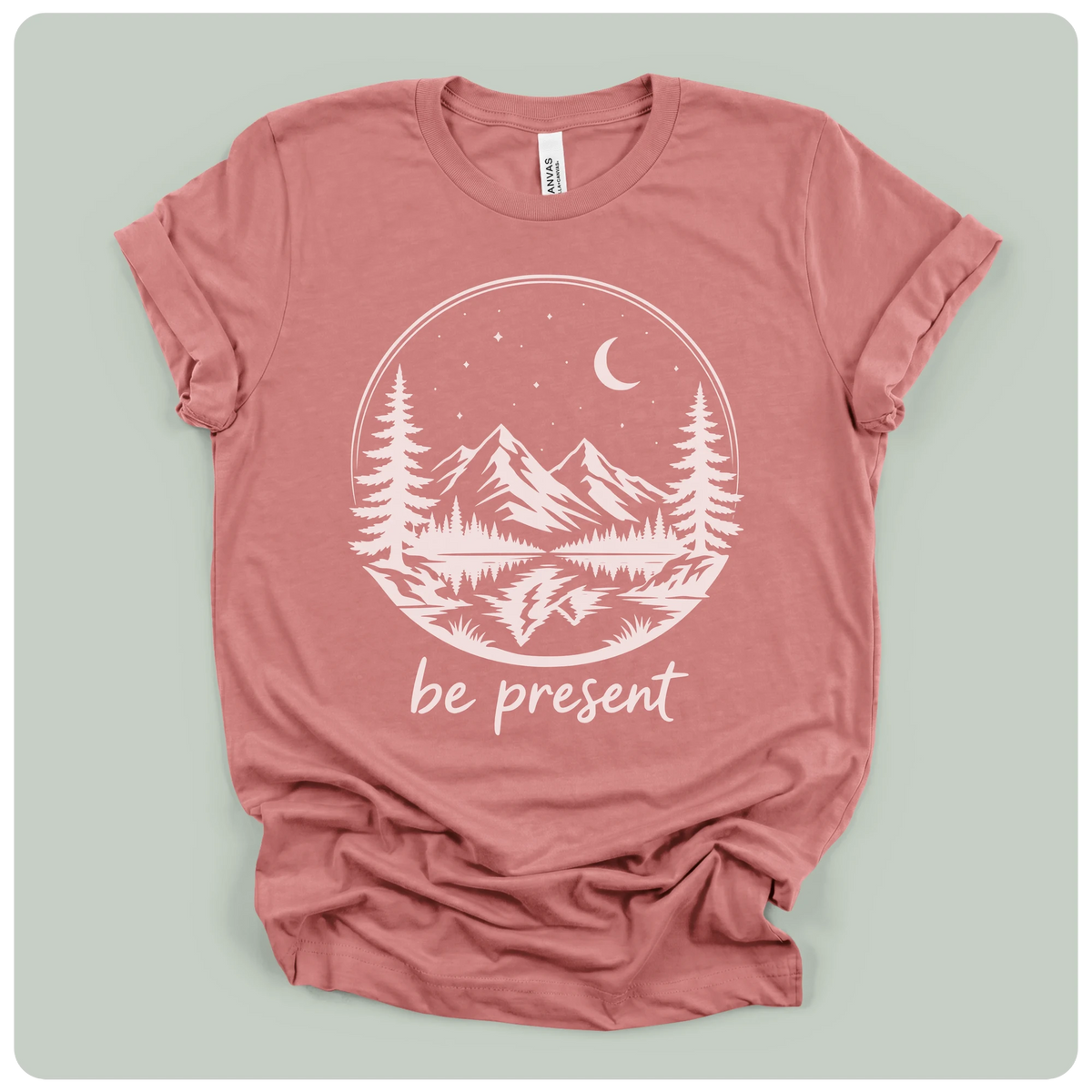 A dusty rose short-sleeve t-shirt with a white graphic print of a mountain landscape at night with the text 'be present' underneath, laid flat on a light sage green background.