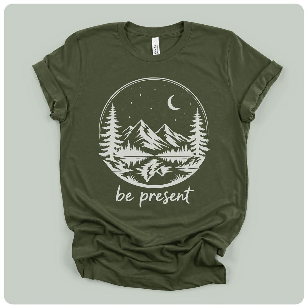 short-sleeve t-shirt with a white graphic print of a mountain landscape at night with the text 'be present' underneath, laid flat on a light sage green background.