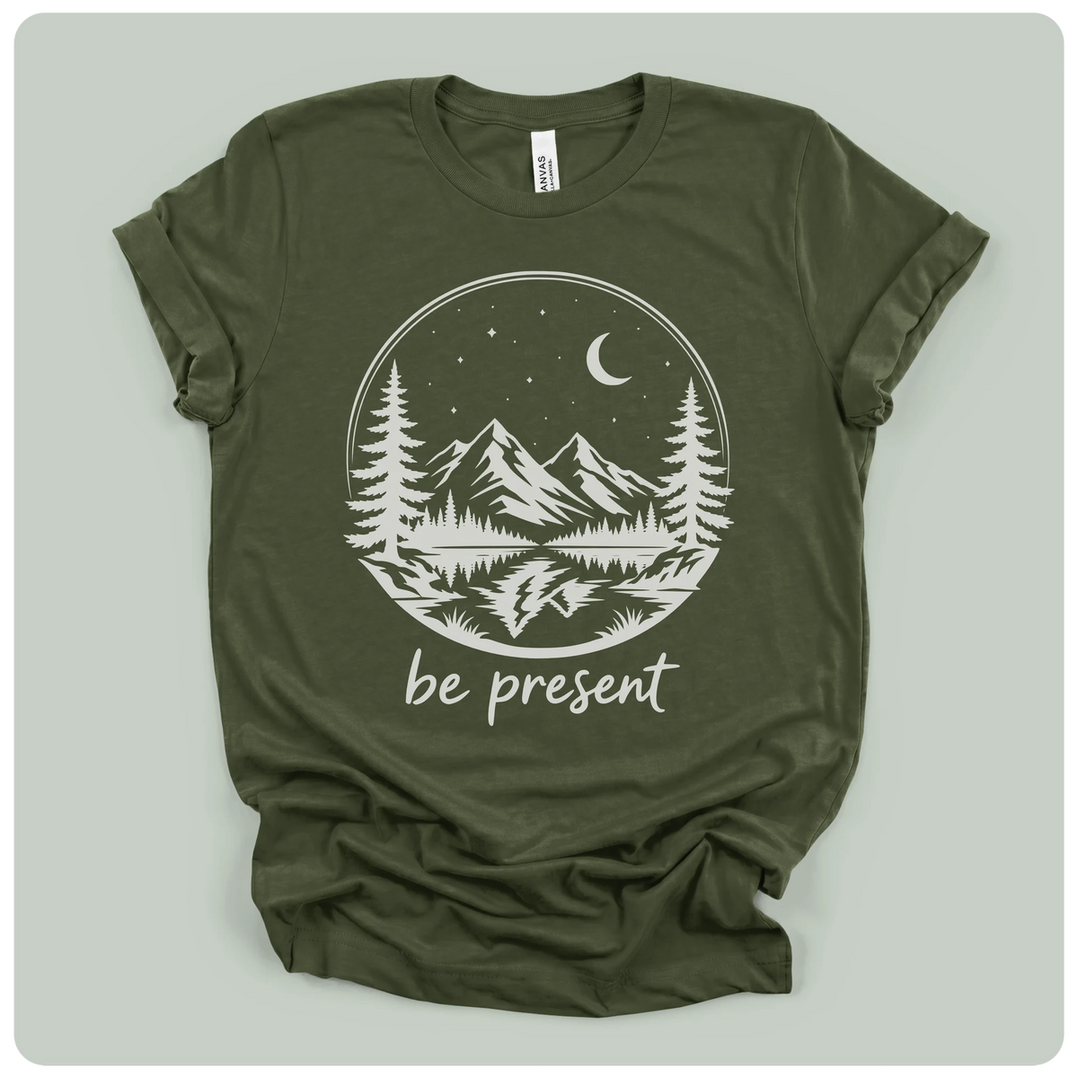 short-sleeve t-shirt with a white graphic print of a mountain landscape at night with the text 'be present' underneath, laid flat on a light sage green background.