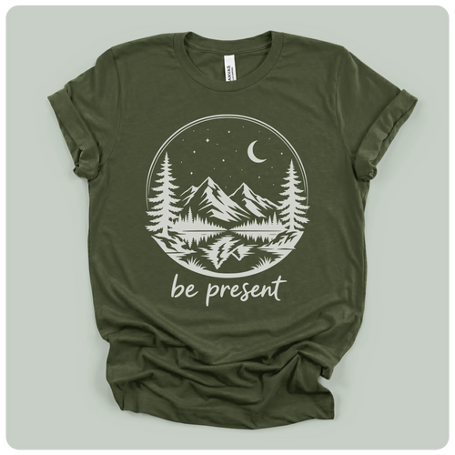 short-sleeve t-shirt with a white graphic print of a mountain landscape at night with the text 'be present' underneath, laid flat on a light sage green background.