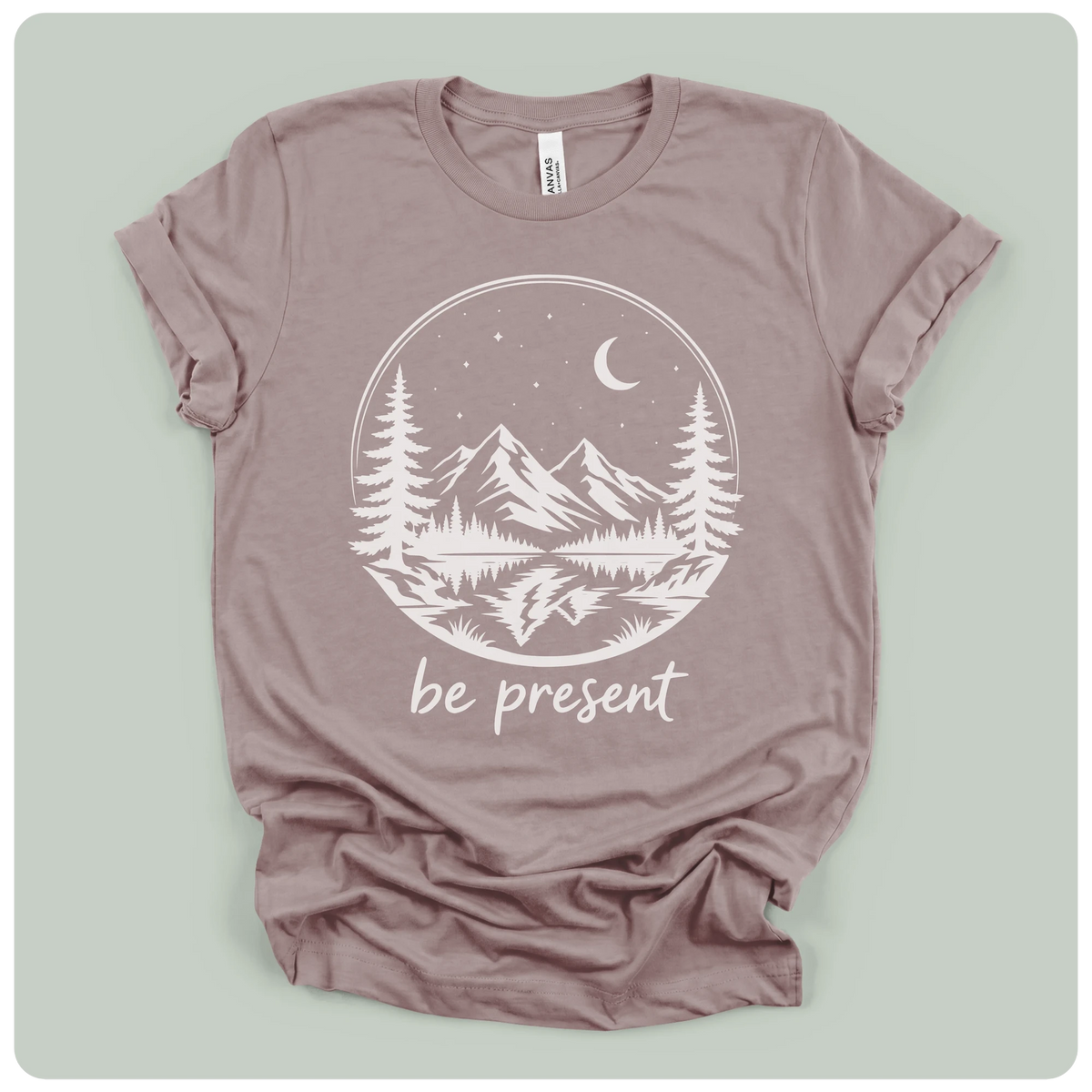 A dusty mauve crewneck t-shirt with a white graphic print of a mountain landscape at night with the text 'be present' underneath, displayed flat on a light sage green background.