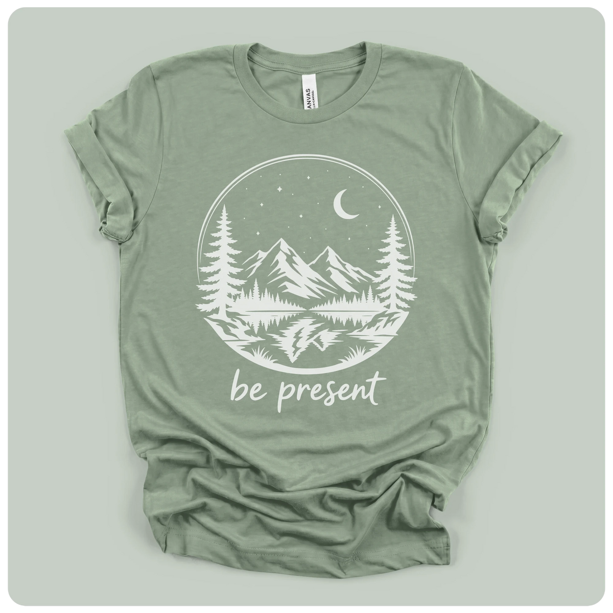 short-sleeve t-shirt with a white graphic print of a mountain landscape at night with the text 'be present' underneath, laid flat on a light sage green background.