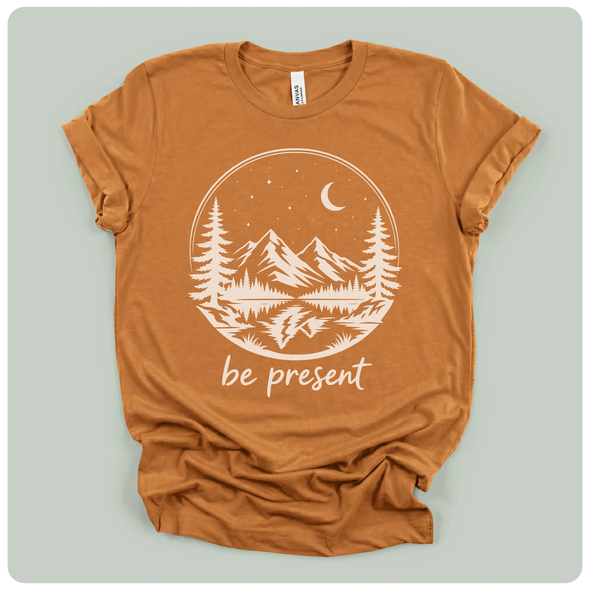 A rust-orange crewneck t-shirt with a white graphic print of a mountain landscape under a crescent moon and stars, with the text 'be present' below, laid flat on a light gray background.