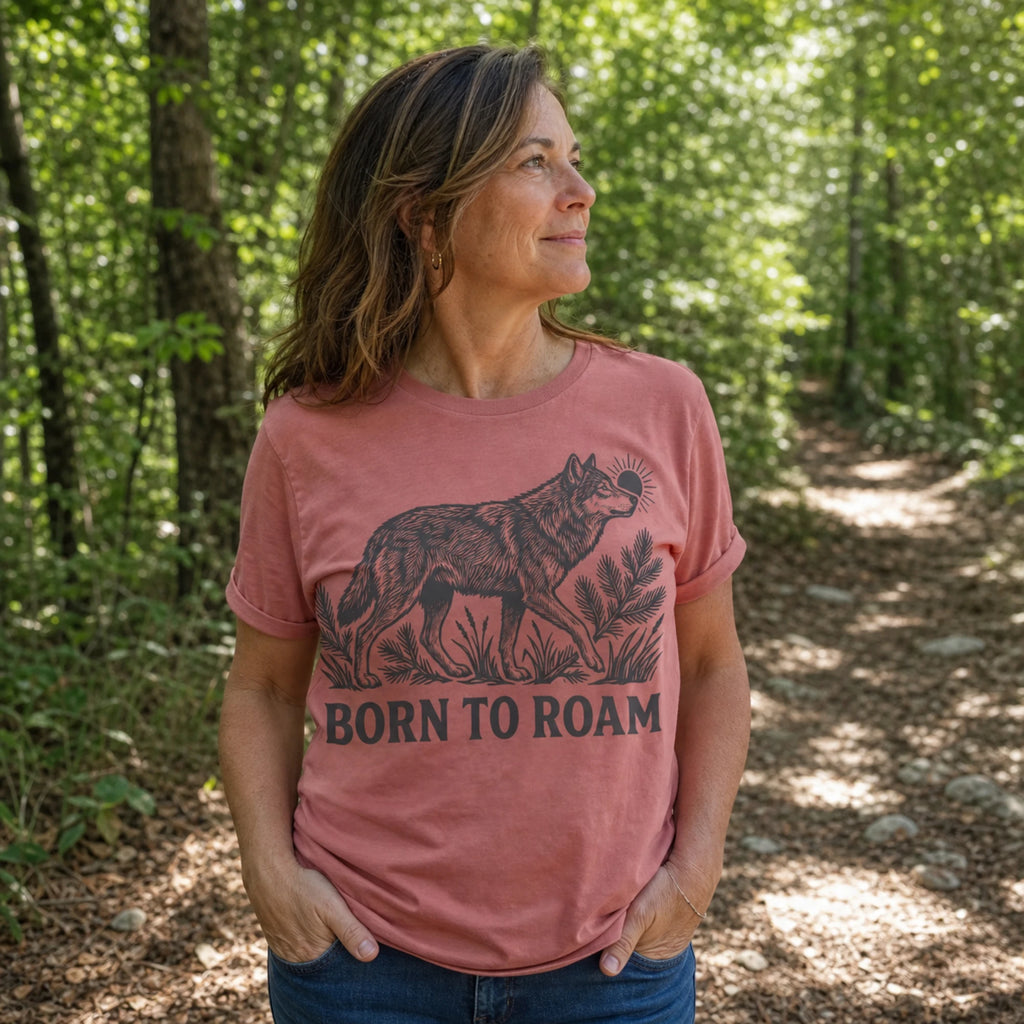A woman standing in a sun-dappled forest wearing a dusty rose pink t-shirt with a black graphic of a wolf and the text 'BORN TO ROAM'.