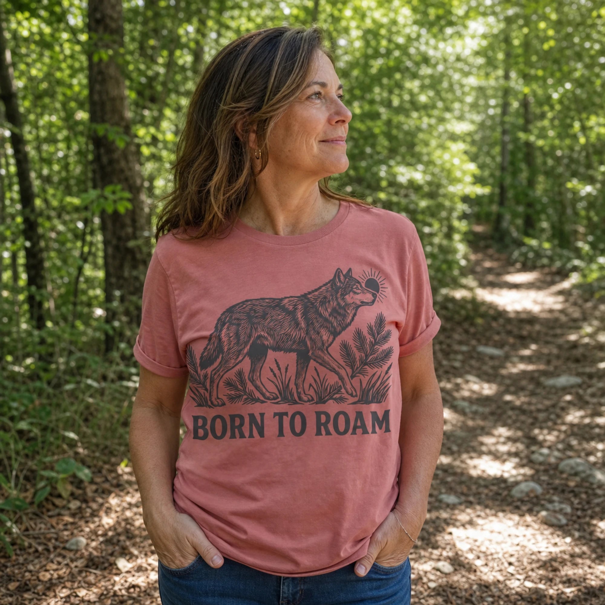 A woman standing in a sun-dappled forest wearing a dusty rose pink t-shirt with a black graphic of a wolf and the text 'BORN TO ROAM'.