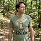 A man wearing an olive green graphic t-shirt with a cartoon groundhog and the text 'STATUS: CURRENTLY UNAVAILABLE' standing in a wooded forest path.