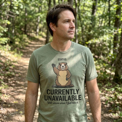 A man wearing an olive green graphic t-shirt with a cartoon groundhog and the text 'STATUS: CURRENTLY UNAVAILABLE' standing in a wooded forest path.