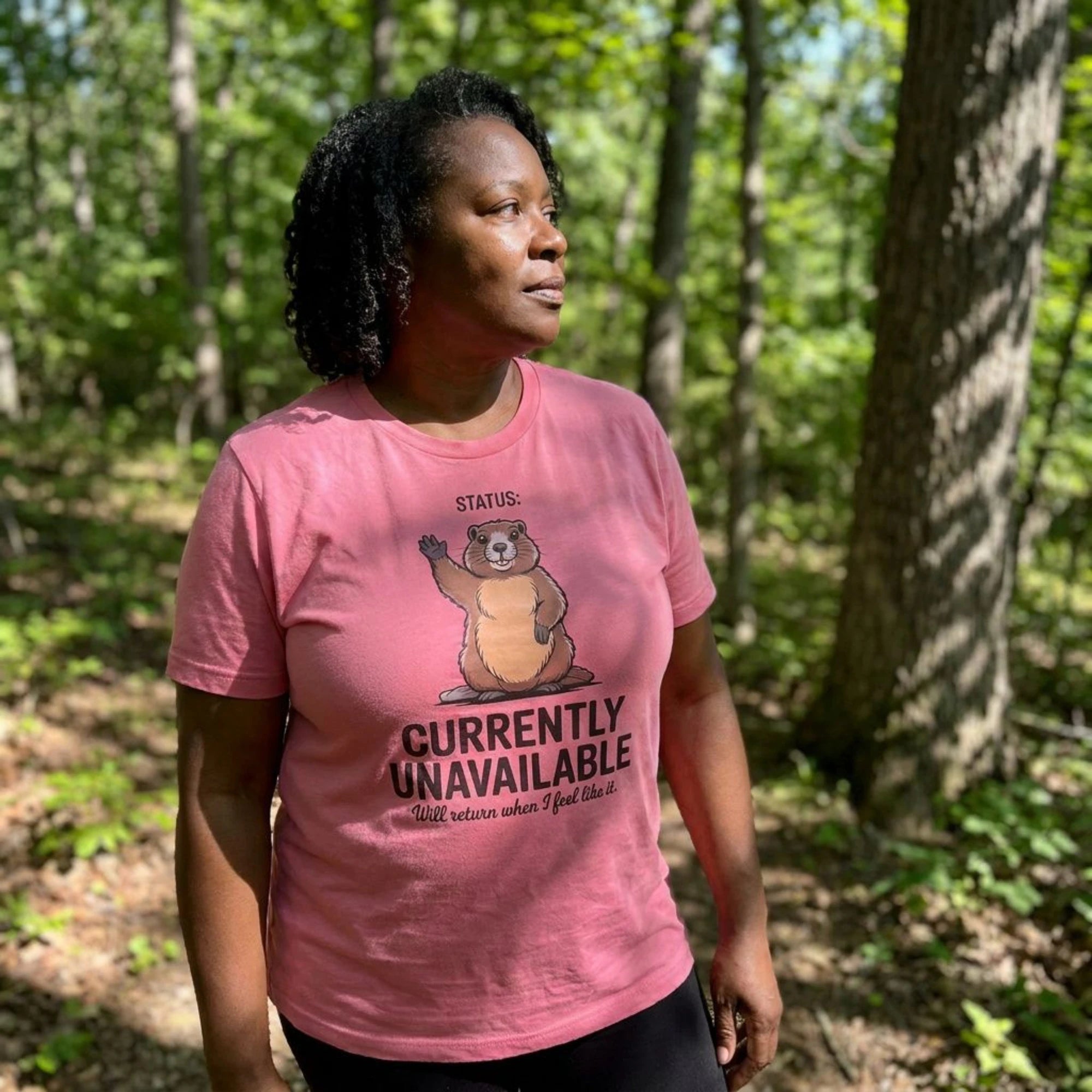 A woman wearing a pink graphic t-shirt with a cartoon groundhog and the text 'STATUS: CURRENTLY UNAVAILABLE' standing in a wooded forest setting.
