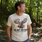 A man wearing a cream-colored t-shirt with a moose graphic and the text 'DO NOT DISTURB Mode Permanently activated.' standing in a wooded forest setting.