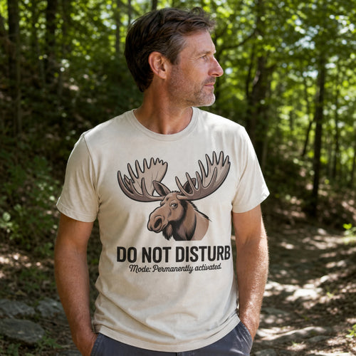 A man wearing a cream-colored t-shirt with a moose graphic and the text 'DO NOT DISTURB Mode Permanently activated.' standing in a wooded forest setting.