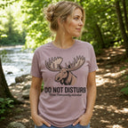 A woman wearing a mauve-colored graphic t-shirt with a moose design and the text 'DO NOT DISTURB' and 'Mode Permanently activated.' standing in a wooded outdoor setting near a river.