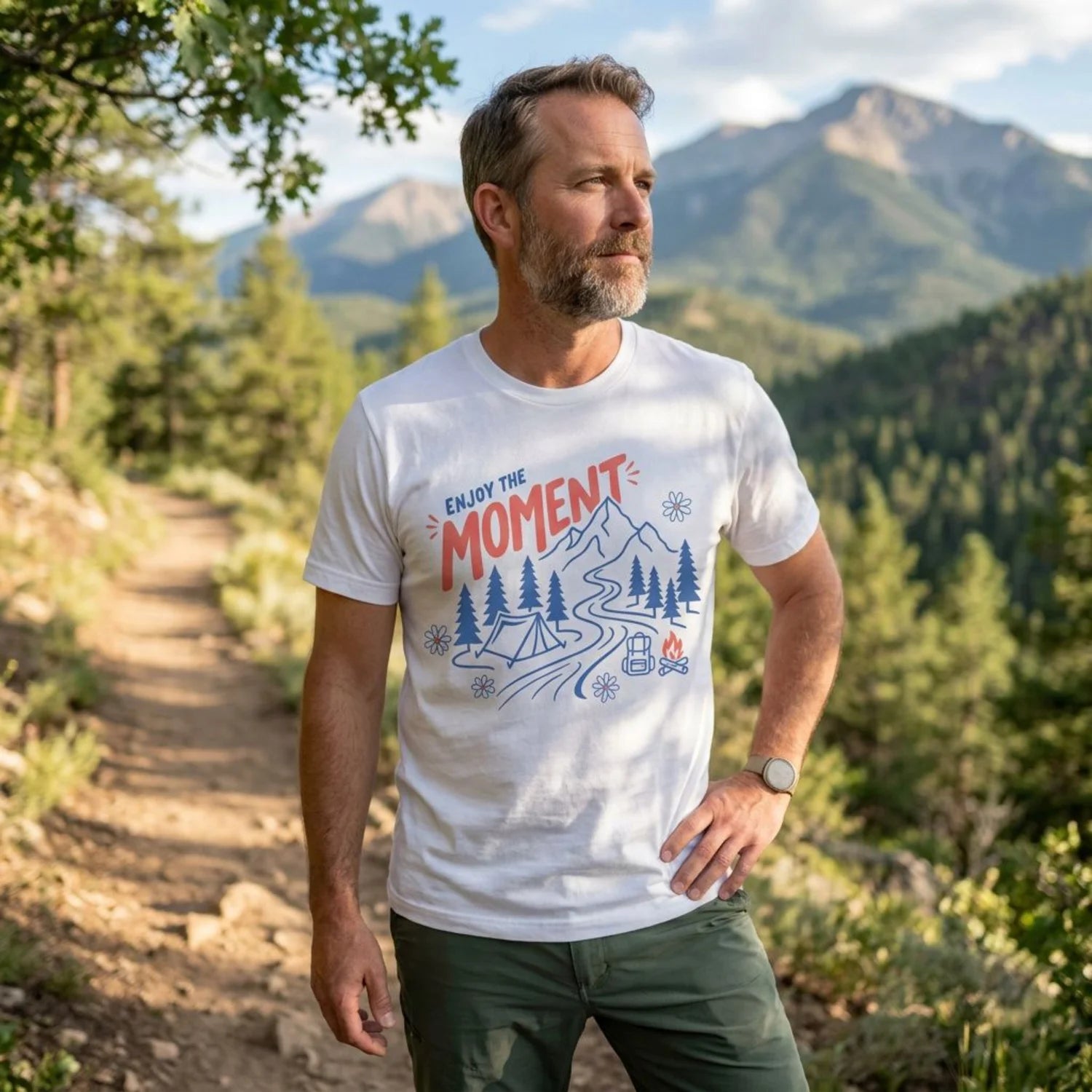 A man standing on a mountain trail wearing a white graphic t-shirt with a blue and red 'ENJOY THE MOMENT' design, paired with dark green pants.