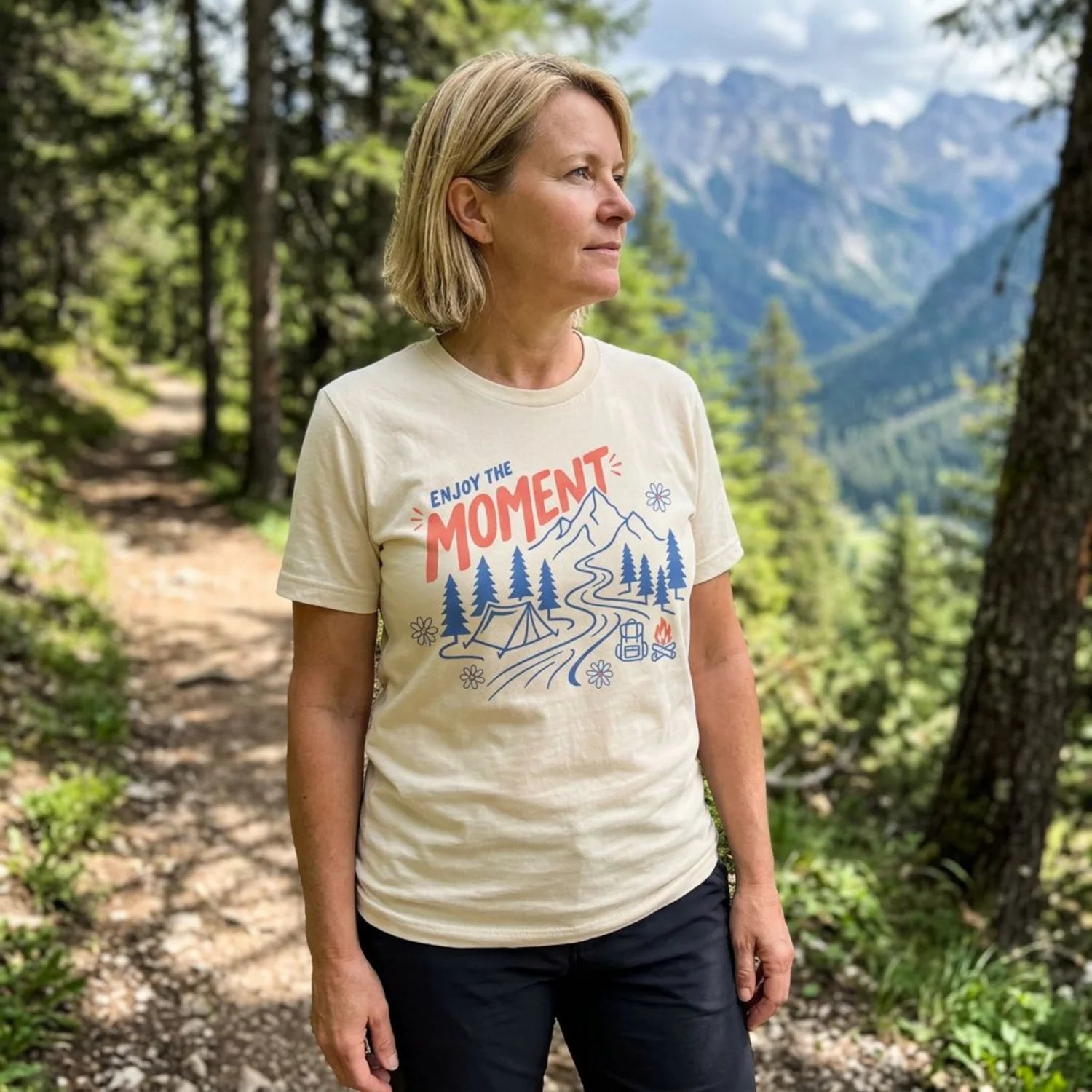 A woman standing on a forest path wearing a beige graphic t-shirt with a blue and red mountain design that reads 'ENJOY THE MOMENT'.