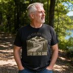 A man wearing a black graphic t-shirt with a mountain road design and the text 'FIND YOUR OWN SIGNAL' and 'STAY OFF THE GRID', standing outdoors in a wooded area near a lake.