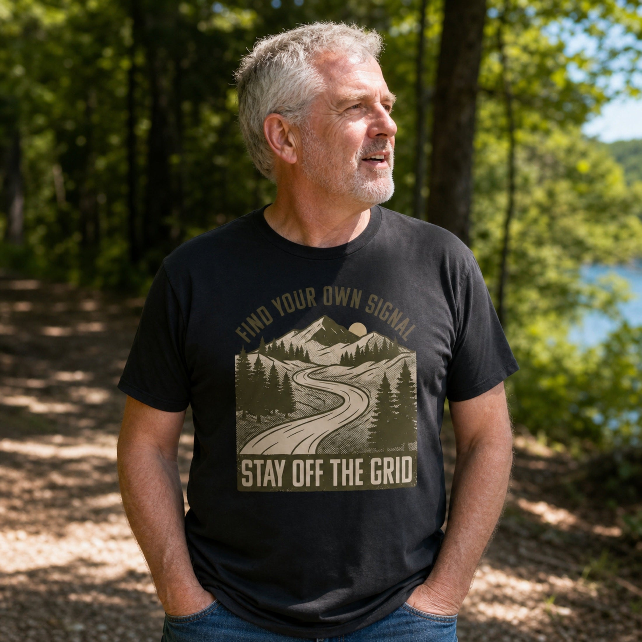 A man wearing a black graphic t-shirt with a mountain road design and the text 'FIND YOUR OWN SIGNAL' and 'STAY OFF THE GRID', standing outdoors in a wooded area near a lake.