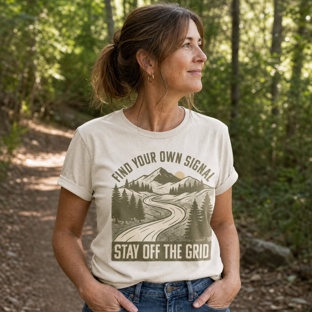 A woman wearing a cream-colored graphic t-shirt with a mountain and road design that reads 'FIND YOUR OWN SIGNAL' and 'STAY OFF THE GRID'. She is standing outdoors on a wooded path.