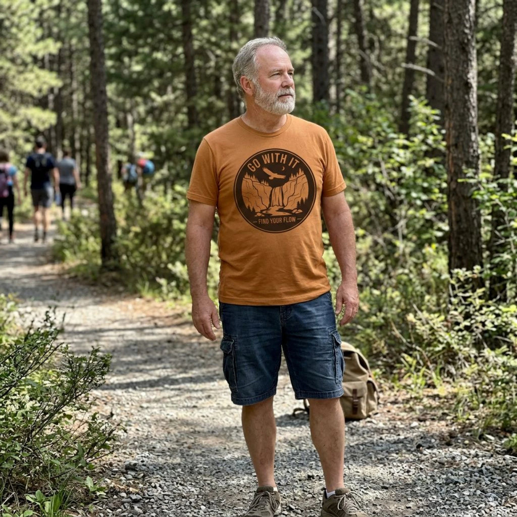 A man hiking on a forest path wearing an orange graphic t-shirt with a black circular logo that reads 'GO WITH THE LOST HUNTER', dark blue cargo shorts, and hiking boots.