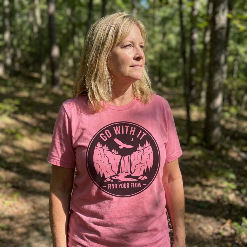 A woman standing in a sunlit forest wearing a pink short-sleeve T-shirt with a large circular black graphic on the chest that reads “GO WITH IT” and “FIND YOUR FLOW.”.