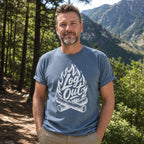 A man wearing a slate blue t-shirt with a white graphic that reads 'Log Out' over crossed logs and flames, standing outdoors in a forest with mountains in the background.