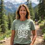 A woman standing on a mountain trail wearing a sage green t-shirt with a white graphic that reads 'Log Out' over a campfire design. The background shows a scenic forest and mountains.