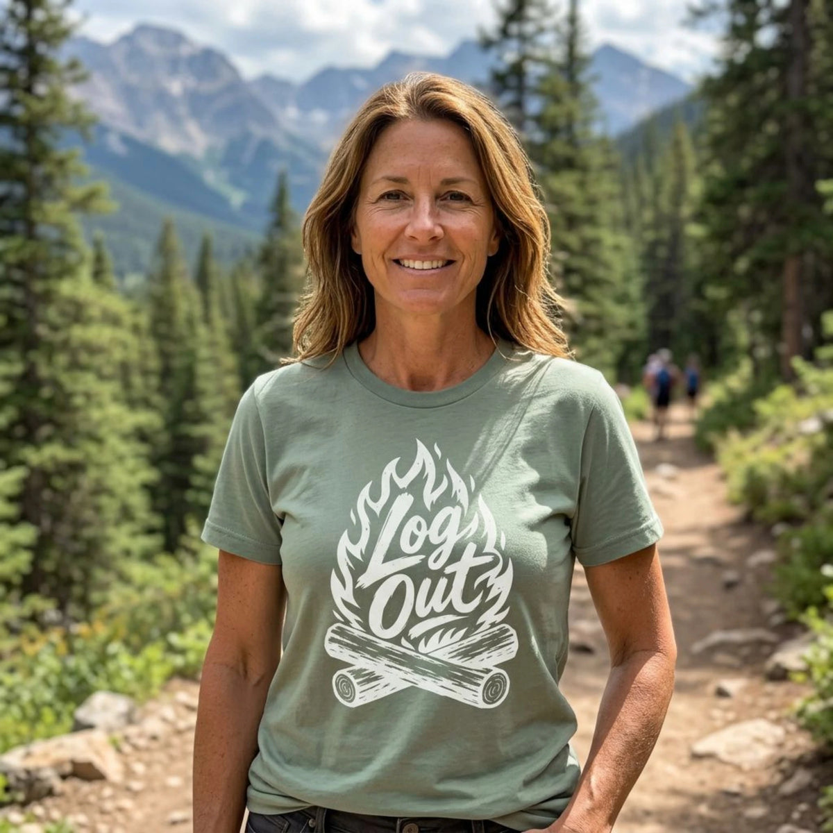 A woman standing on a mountain trail wearing a sage green t-shirt with a white graphic that reads 'Log Out' over a campfire design. The background shows a scenic forest and mountains.