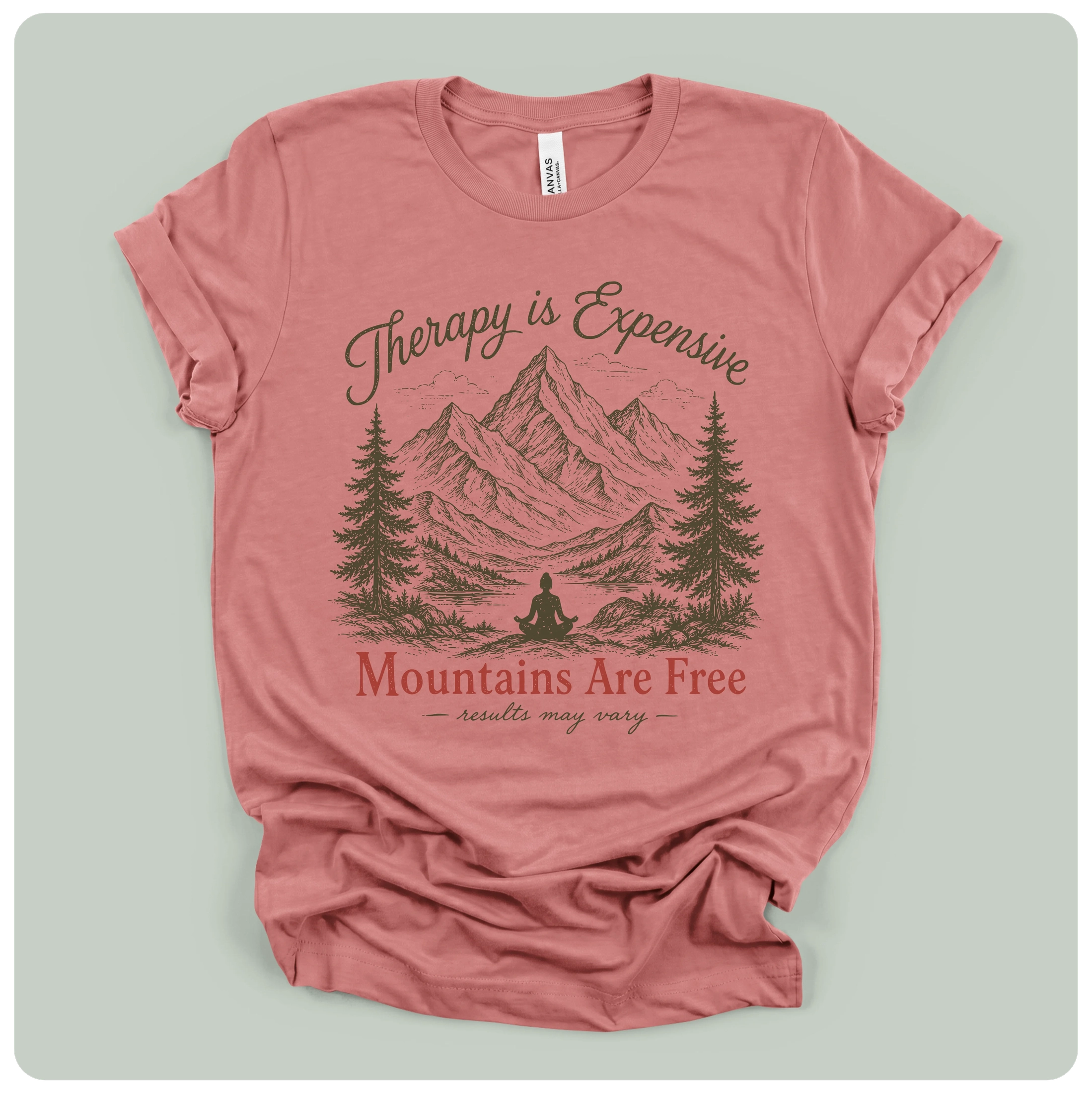 A dusty rose pink t-shirt with a graphic print of mountains, pine trees, and a meditating figure, with the text 'Therapy is Expensive' and 'Mountains Are Free'.
