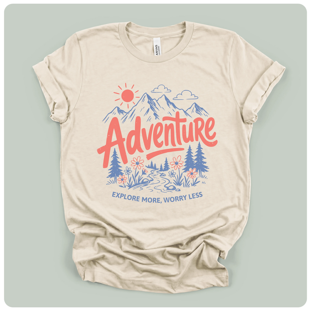 A beige short-sleeve t-shirt with a colorful graphic print on the front. The graphic features a red sun, blue mountains, pine trees, and the word 'Adventure' in red script, with the phrase 'EXPLORE MORE, WORRY LESS' printed below in blue.