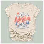 A beige short-sleeve t-shirt with a colorful graphic print on the front. The graphic features a red sun, blue mountains, pine trees, and the word 'Adventure' in red script, with the phrase 'EXPLORE MORE, WORRY LESS' printed below in blue.