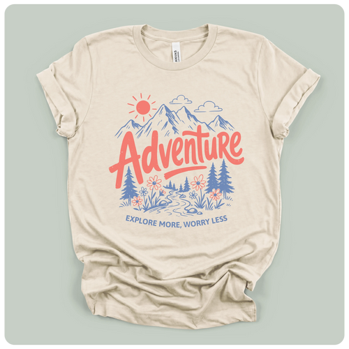A beige short-sleeve t-shirt with a colorful graphic print on the front. The graphic features a red sun, blue mountains, pine trees, and the word 'Adventure' in red script, with the phrase 'EXPLORE MORE, WORRY LESS' printed below in blue.