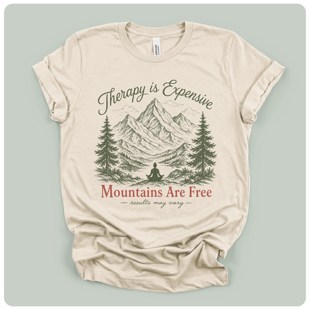 A beige short-sleeve t-shirt with a graphic print of mountains, pine trees, and a meditating figure, with the text 'Therapy is Expensive' and 'Mountains Are Free'.