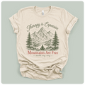 A beige short-sleeve t-shirt with a graphic print of mountains, pine trees, and a meditating figure, with the text 'Therapy is Expensive' and 'Mountains Are Free'.