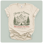 A beige short-sleeve t-shirt with a graphic print of mountains, pine trees, and a meditating figure, with the text 'Therapy is Expensive' and 'Mountains Are Free'.