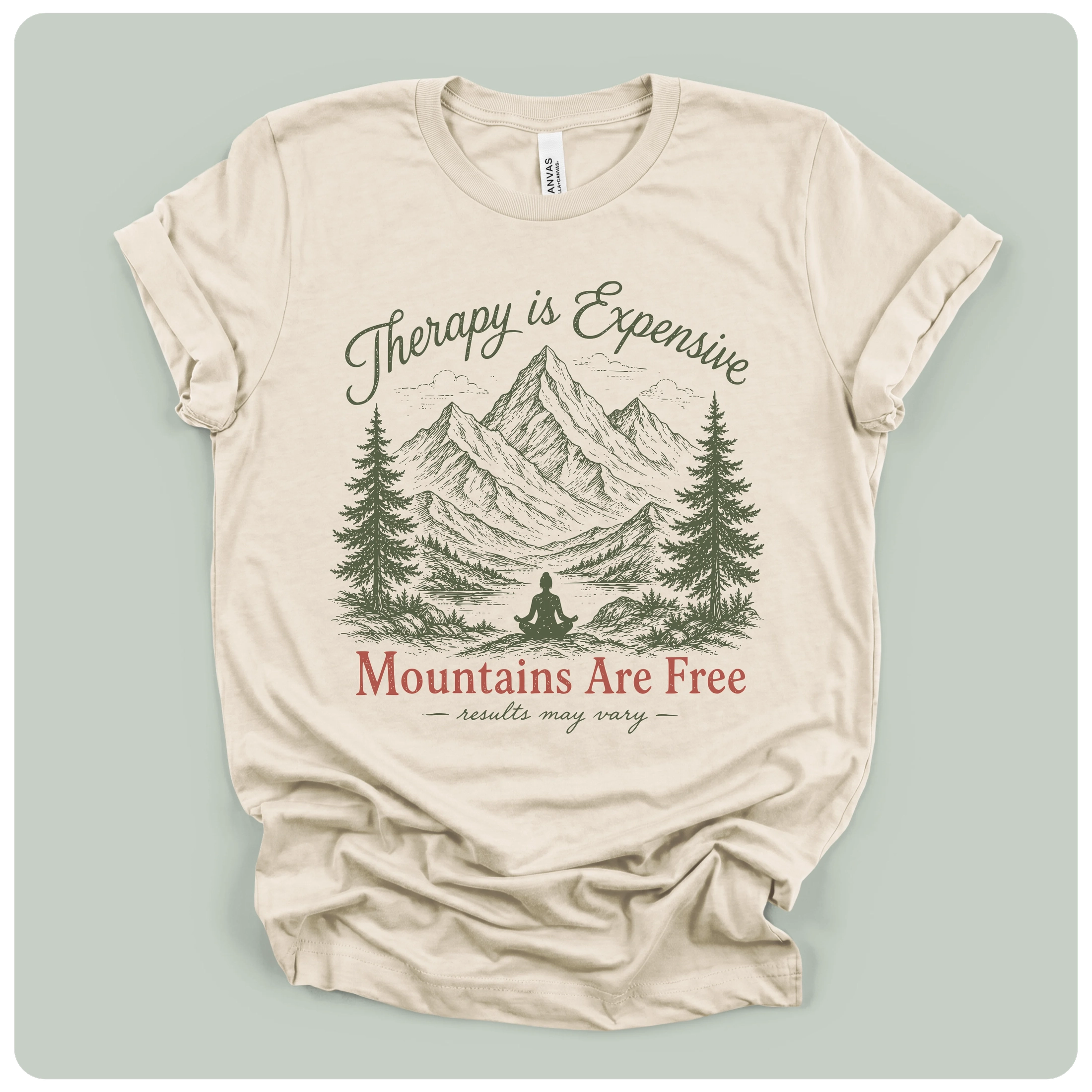 A beige short-sleeve t-shirt with a graphic print of mountains, pine trees, and a meditating figure, with the text 'Therapy is Expensive' and 'Mountains Are Free'.