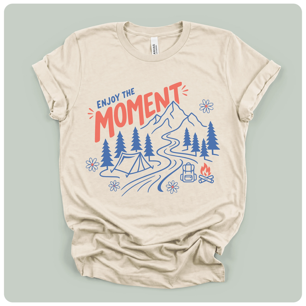 A beige short-sleeve t-shirt with a graphic print of a mountain, campsite, trees, and the text 'ENJOY THE MOMENT' in red and blue, displayed on a light sage green background.