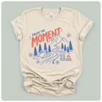 A beige short-sleeve t-shirt with a graphic print of a mountain, campsite, trees, and the text 'ENJOY THE MOMENT' in red and blue, displayed on a light sage green background.