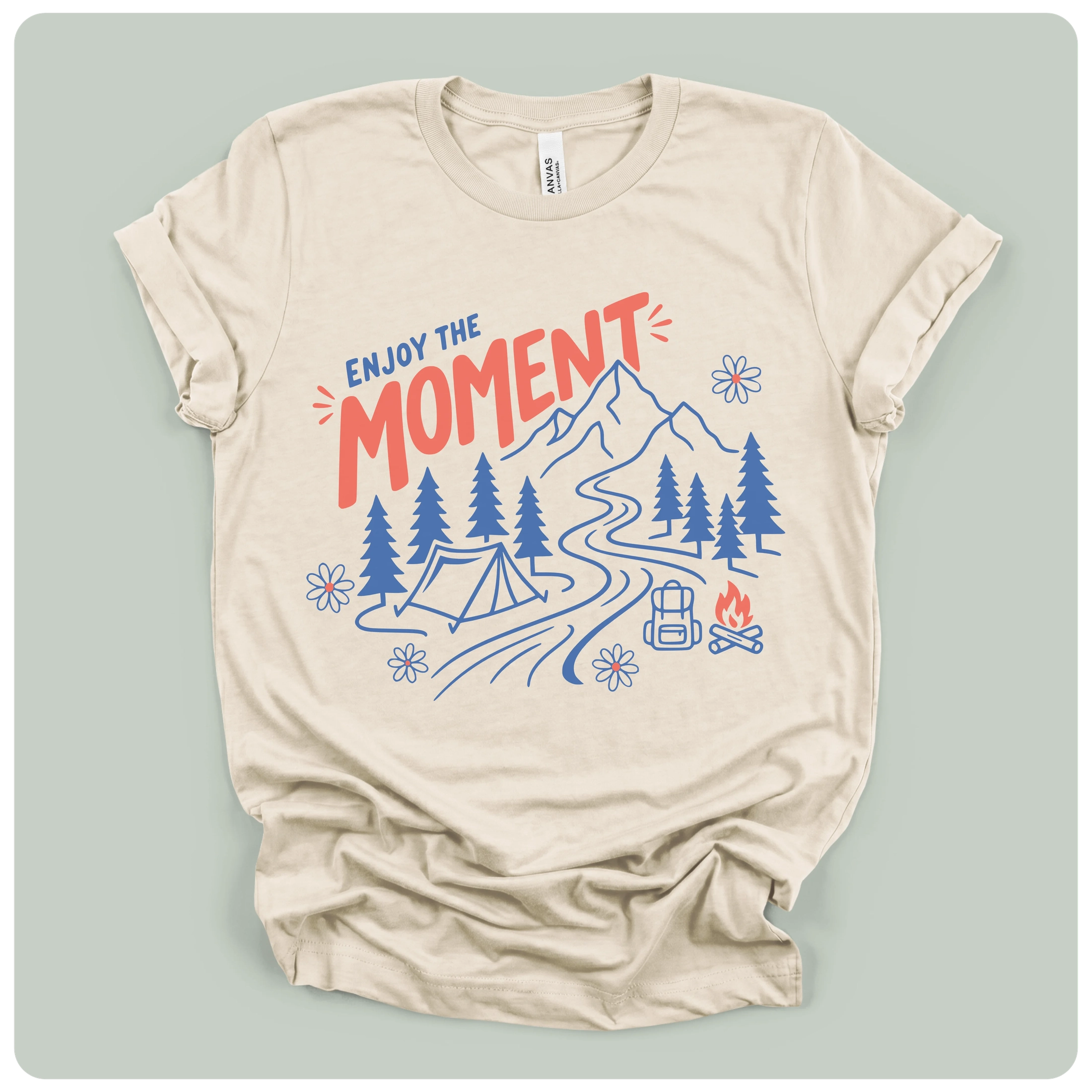A beige short-sleeve t-shirt with a graphic print of a mountain, campsite, trees, and the text 'ENJOY THE MOMENT' in red and blue, displayed on a light sage green background.