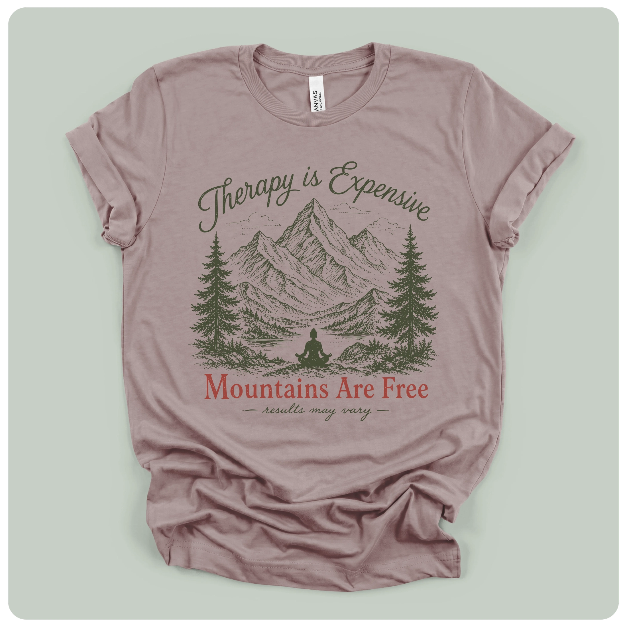 A dusty mauve short-sleeve t-shirt with a graphic print of mountains, pine trees, and a meditating figure. The text reads 'Therapy is Expensive' in dark green script, 'Mountains Are Free' in red serif font, and 'Assaults may vary' in dark green script below.