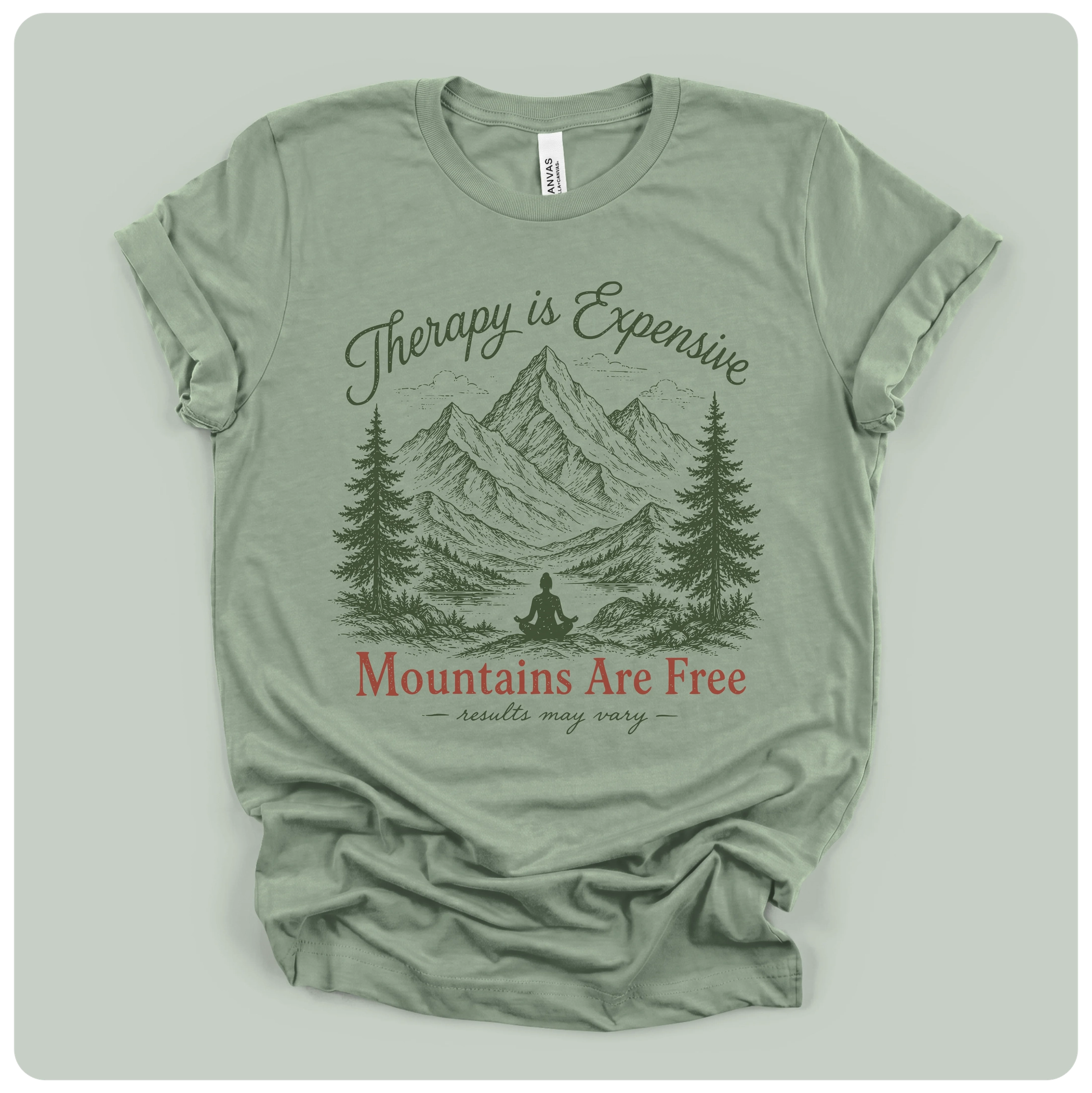 A sage green graphic t-shirt laid flat on a light gray background. The shirt features a dark green line art illustration of mountains, pine trees, and a person meditating, with the text 'Therapy is Expensive' in dark green script and 'Mountains Are Free' in red serif font.