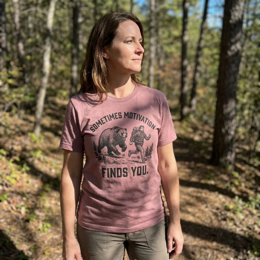 A woman wearing a dusty rose graphic t-shirt with a bear design and the text 'SOMETIMES MOTIVATION FINDS YOU.' standing in a sunlit forest.