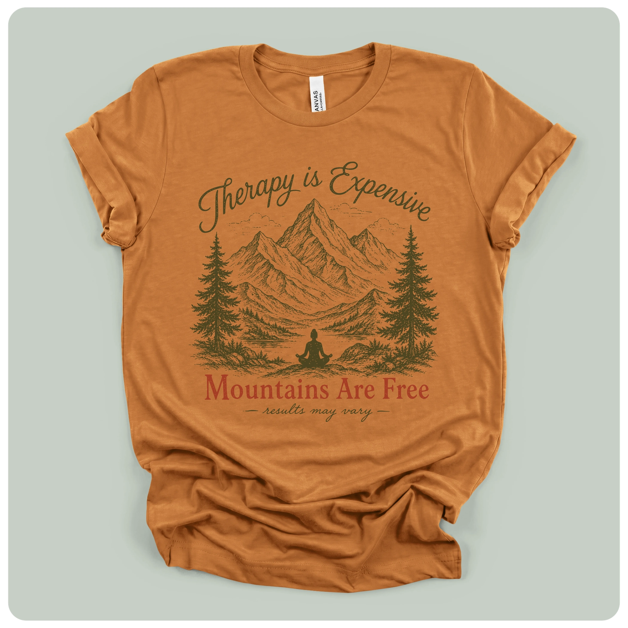 A rust-orange crewneck t-shirt with a distressed graphic of mountains, pine trees, and a meditating figure, with the text 'Therapy is Expensive' and 'Mountains Are Free'.