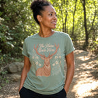 A woman wearing a sage green graphic t-shirt with a deer and the text 'The Noise Ends Here' stands on a sunlit forest path.