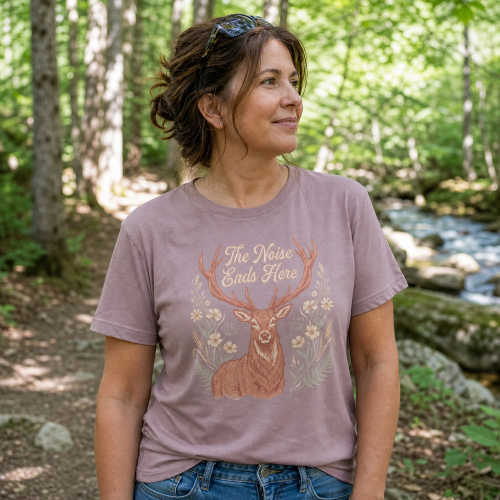 A woman wearing a mauve-colored graphic t-shirt with a deer design and the text 'The Noise Ends Here', standing in a forest with a stream in the background.