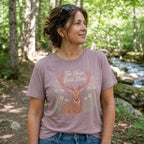 A woman wearing a mauve-colored graphic t-shirt with a deer design and the text 'The Noise Ends Here', standing in a forest with a stream in the background.